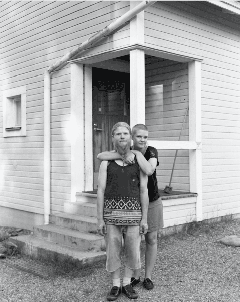 Black and white photograph of a young couple embraced standing in front of their wooden clad home.